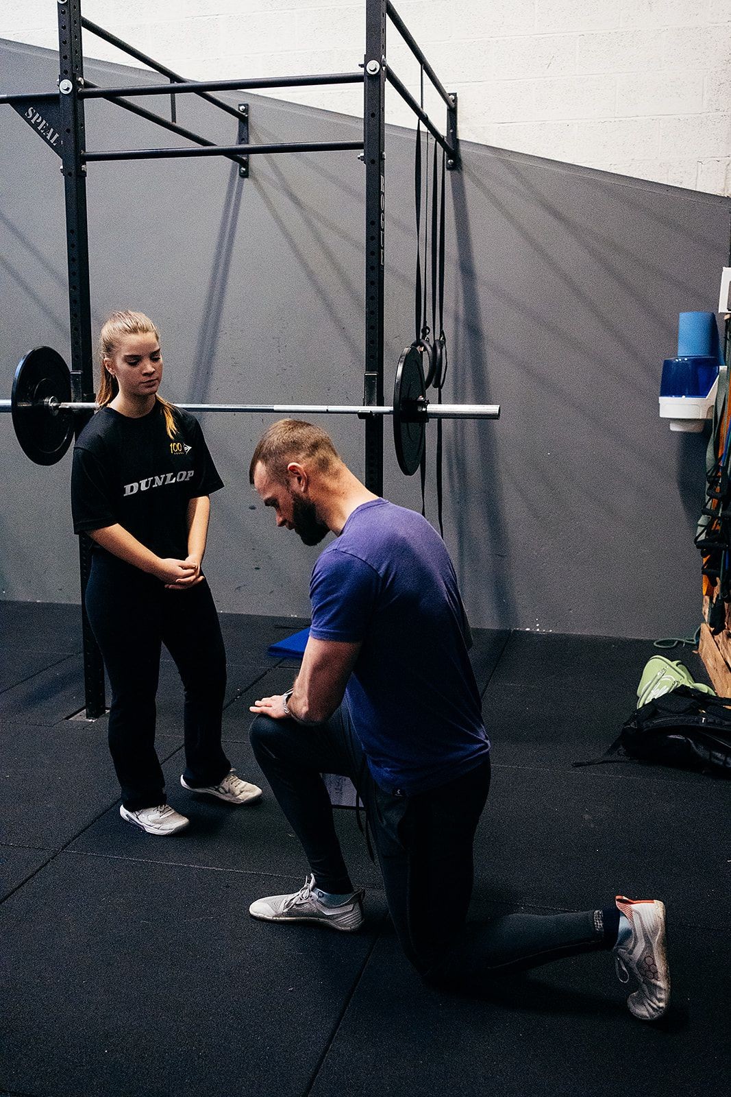 Man kneeling and woman standing in a gym with a barbell and rack in the background.