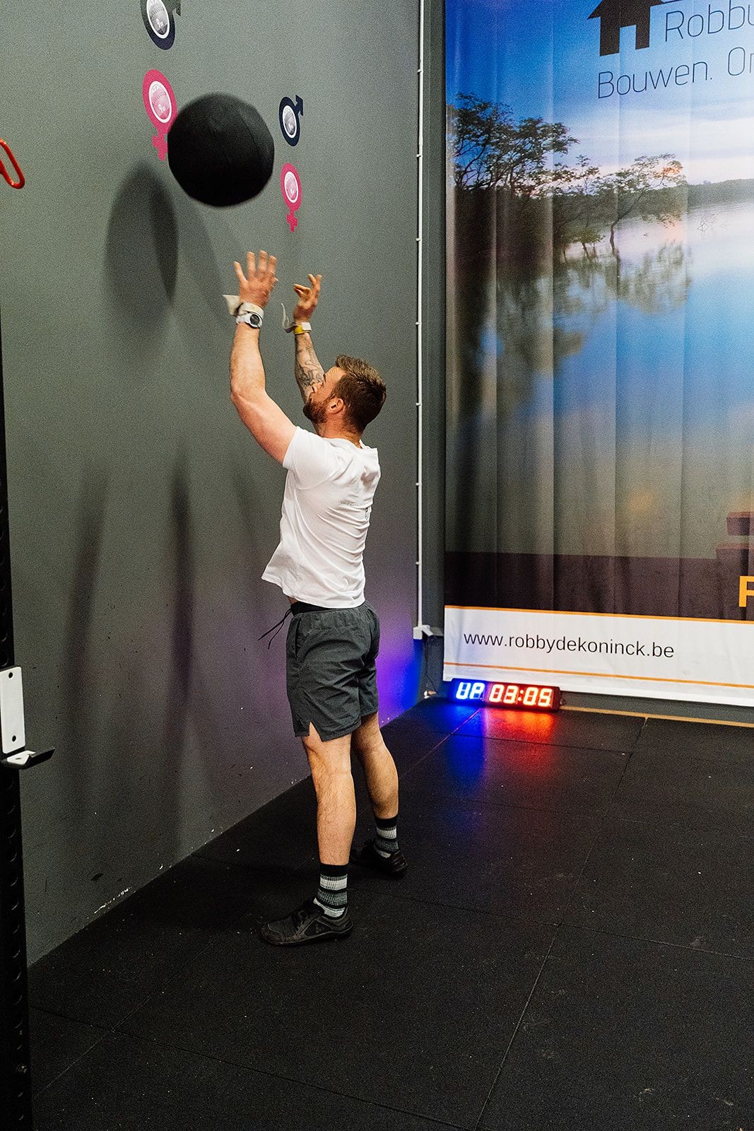 Man performing a wall ball exercise in a gym with timer and logo visible.