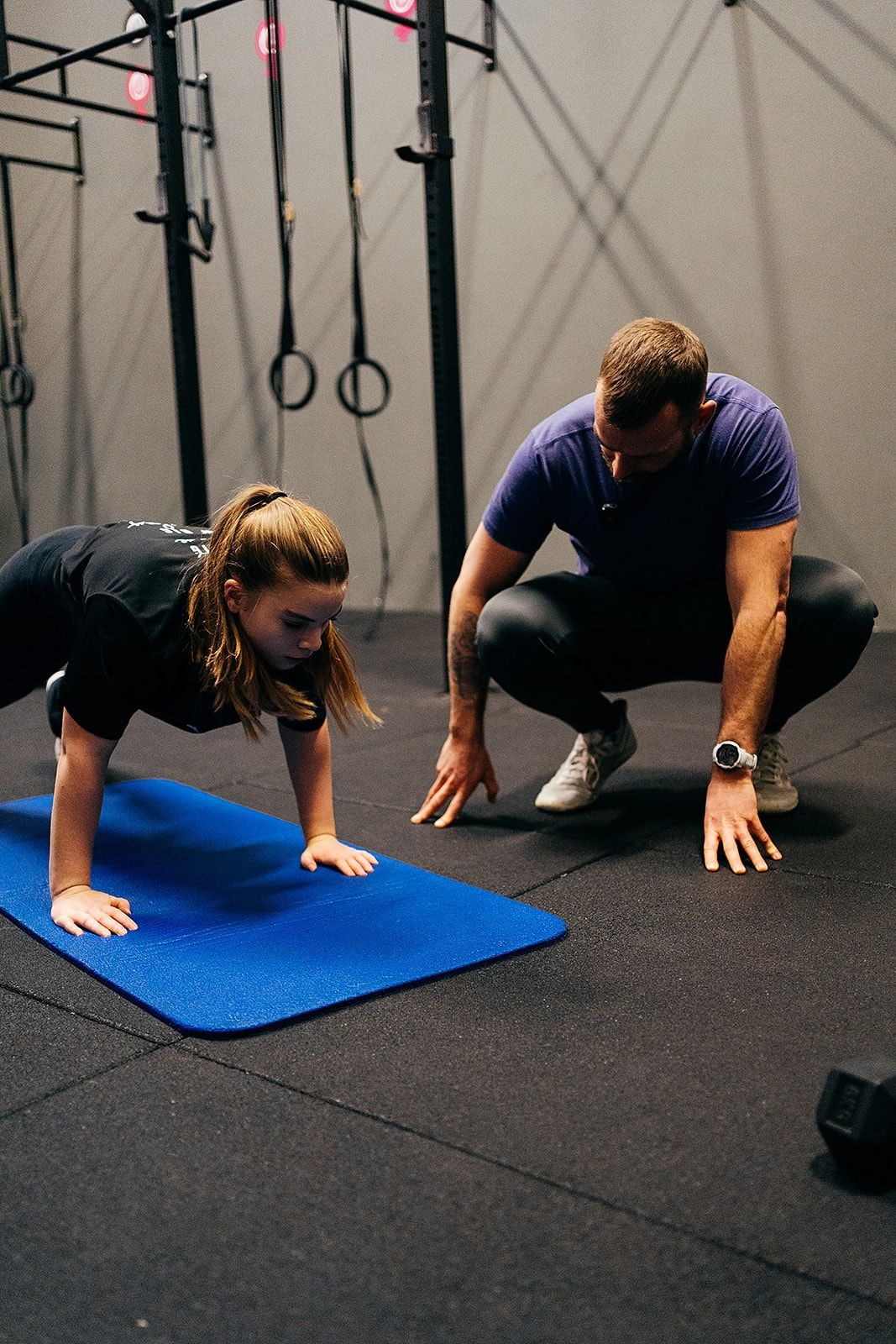Instructor guiding a young girl doing a push-up on a blue mat in a gym setting.