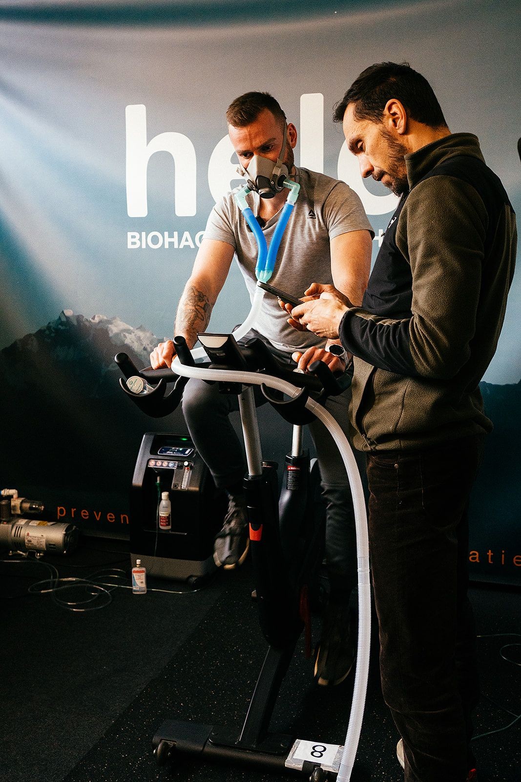 Man on exercise bike wearing a mask connected to tubes, with another man using a phone nearby.