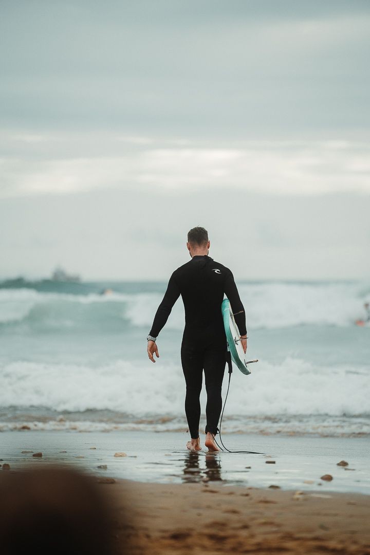 Surfer in wetsuit walking towards the ocean with a surfboard on a cloudy day.
