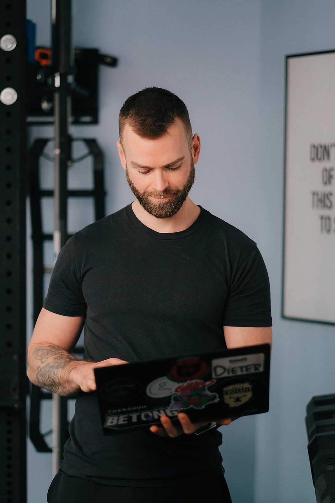 Man with a beard wearing a black t-shirt using a laptop, standing indoors.